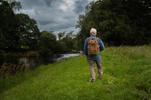Mature man standing beside a river in a rural location in Scotland on an overcast summer afternoon