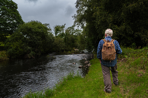 Senior man standing beside a river in a rural location in Scotland on an overcast summer afternoon