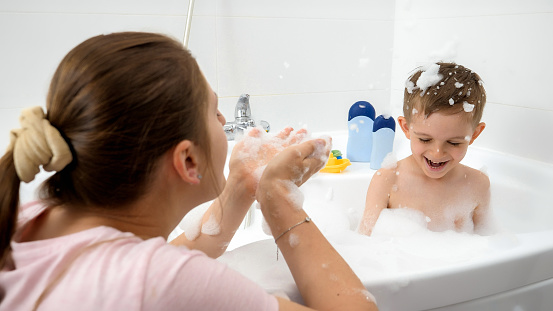 Cheerful smiling mother with son playing and having fun inbath full of soap foam and suds.