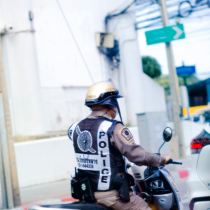 Capture of Thai policeman on motorcycle waiting in queued traffic at traffic light on motorcycle. Officer is wearing golden colored crash helmet and uniform i including gun. Scene is in Bangkok between Ramintra and Lat Krabang