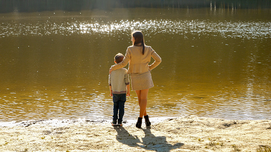 Happy boy with mother standing on riverbank at sunset rays. Family outdoors, happy parenting and childhood, autumn landscape, kids and parents relaxing outside