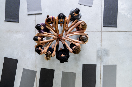 A large group of adults are seen standing in a circle with their hands stacked in the middle as they cheer and celebrate after a workout.  They are each dressed in athletic wear in this aerial view.