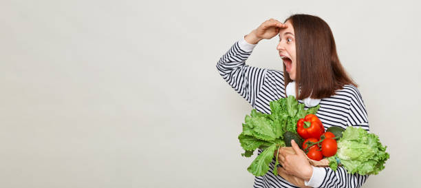 Surprised woman wearing striped casual shirt holding lettuce cabbage pepper avocado isolated over gray background looking far at mockup for advertisement area.