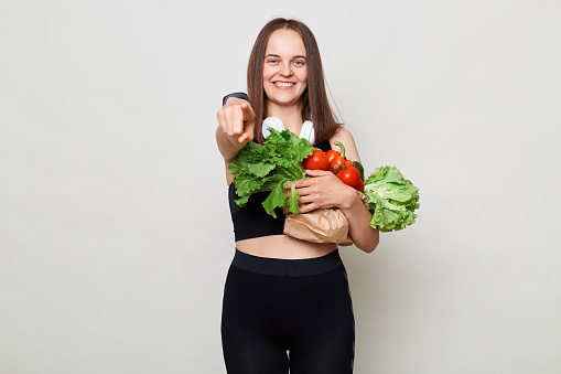 Smiling joyful woman wearing sportswear carrying vegetable isolated over gray background pointing to camera choosing inviting you to do sport and eat healthy food.