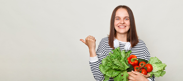 Cheerful woman wearing striped casual shirt carrying vegetable isolated over gray background pointing at copy space for advertisement mockup.