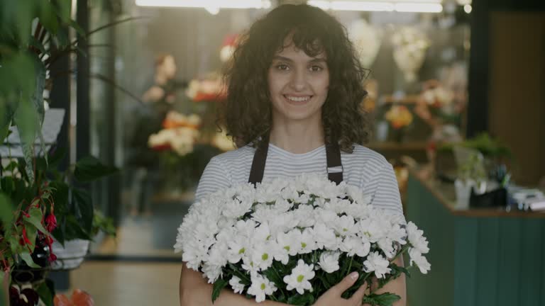 Portrait of cheerful saleswoman in uniform standing in flower shop holding beautiful bouquet and looking at camera
