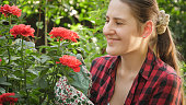 Portrait of happy smiling female gardener looking and smelling blooming red roses in garden. Gardening hobby and plant growing