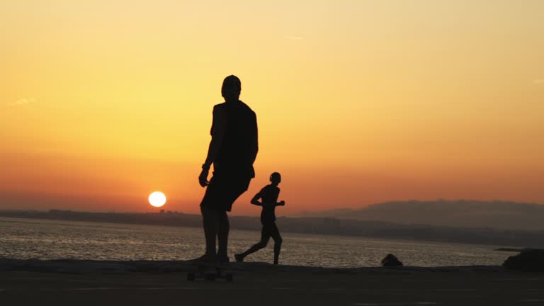 Silhouette of a man skating on a longboard at bright orange sunset by the ocean