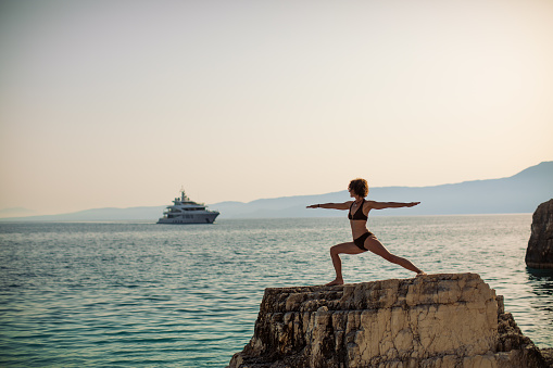 Beautiful fit and muscular mature woman doing yoga on top of  the rock in sea. Calm morning and beautiful sea