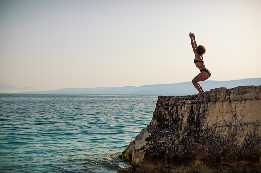 Beautiful fit and muscular mature woman doing yoga on top of  the rock in sea. Calm morning and beautiful sea