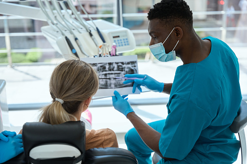 A young african american dentist, technician, or dental assistant places a modern digital x-ray device on the jaw of a female patient to determine the health of her teeth.