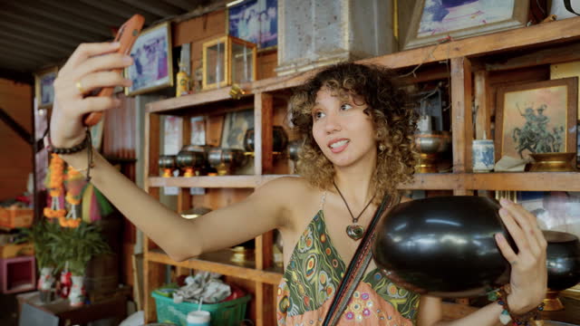 Happy young woman taking a selfie with a monk's bowl at a handicraft shop in Bangkok, Thailand.