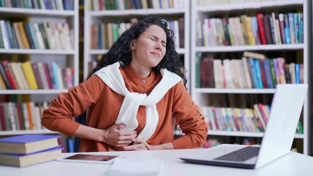 Sad female student suffering from stomach pain while working on laptop in campus library space