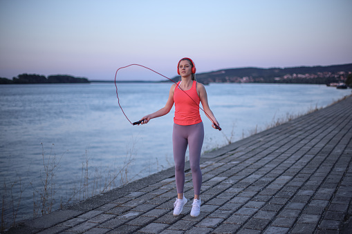 Beautiful young girl in her 20s with blonde hair by the river on a fitness path exercising with jumping rope while listening to music and enjoying the sunset. Photo taken in the golden hour