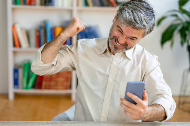 istock Excited overjoyed young man sitting at table holding smartphone, feeling euphoria from mobile online bet gambling. Enthusiastic guy looking at mobile phone screaming with joy, celebrating receiving fantastic news 1612473064