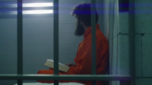 Male prisoner in orange uniform sits on the bed, reads Bible