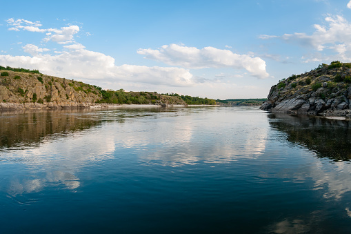 Large cumulus clouds are reflected in the water of a wide river with high rocky banks in the forest and a long beach in the background