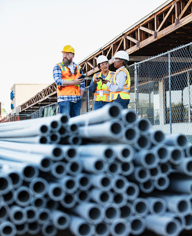 A multiracial group of three construction workers, two women and one man, having a discussion at a job site, a stack of pvc pipes out of focus in the foreground. The project is the renovation of a strip mall.