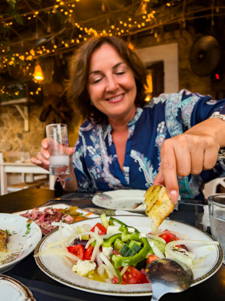 Cheerful woman enjoying food and drink Beautiful middle age woman sitting at a table in a restaurant (taverna) and enjoying wonderful and delicious Mediterranean food with a glass of ouzo, an anis tasted spirit. She's putting her bread in the salasd. Happy female person. mediterranean woman portrait stock pictures, royalty-free photos & images