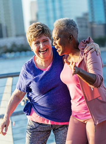 Two multiracial senior women walking together on a city waterfront, conversing and laughing. The woman on the left is Hispanic. Her friend in pink is African-American. They are in their 70s.