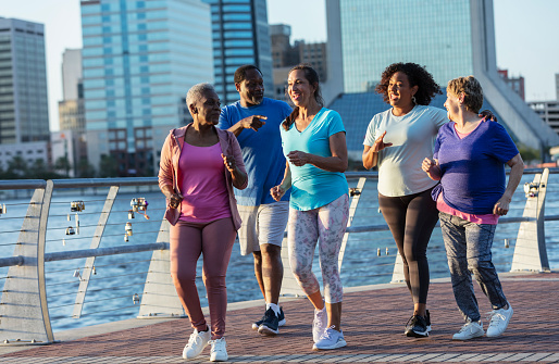 A multiracial group of five mature and senior adults, including one man, exercising on a city waterfront, powerwalking together. They range in age from 40s to 70s.