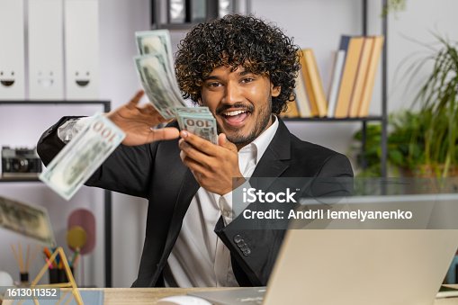 istock Cheerful rich business man working on office laptop wasting throwing money to camera profit win 1613011352