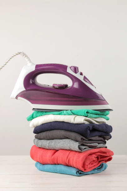 istock Iron and a stack of colored clothes on a white background. Ironing of clothes after washing and cleaning. 1613018192
