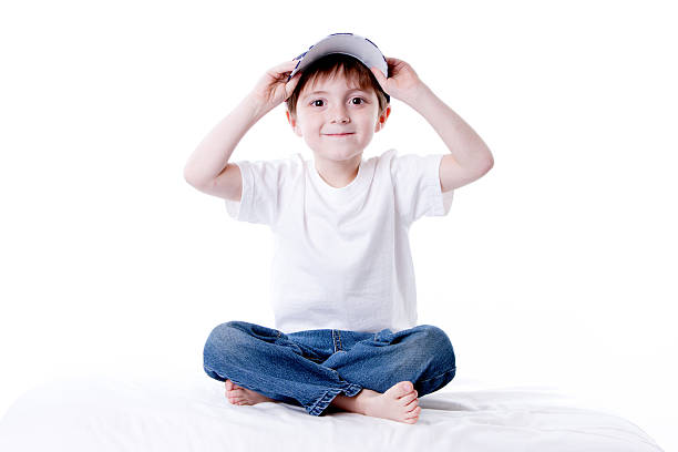 A cheerful young boy outdoors, wearing a bright blue cap and gray long-sleeve shirt, smiles as he adjusts his hat. Bright natural light and a soft green background evoke playful outdoor moments.