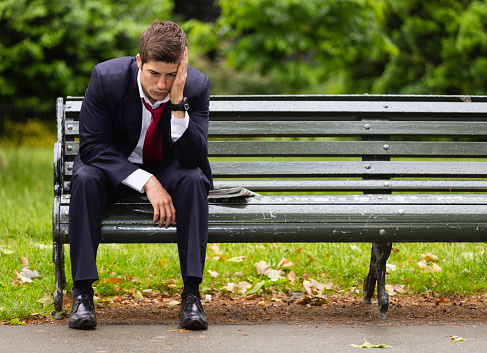 A tired looking businessman sitting alone on a park bench in London on a rainy day.