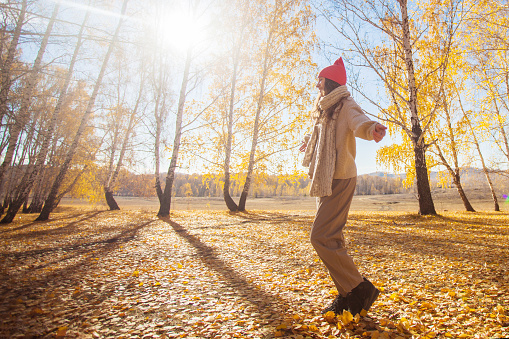 Happy young millennial woman enjoys walking in autumn forest. Teenage girl dressed in warm sweater and hat walks on a sunny day in fall