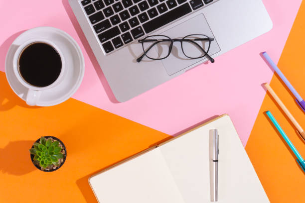 istock Trendy home office workspace with a cup of coffee, a laptop, some pens, a notebook, a pair of glaasses, an artificial plant on pink and orange background 1613564221