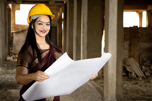 Young beautiful Indian female civil engineer or architect wearing saree and helmet holding paperwork blueprint at construction site. Looking at camera