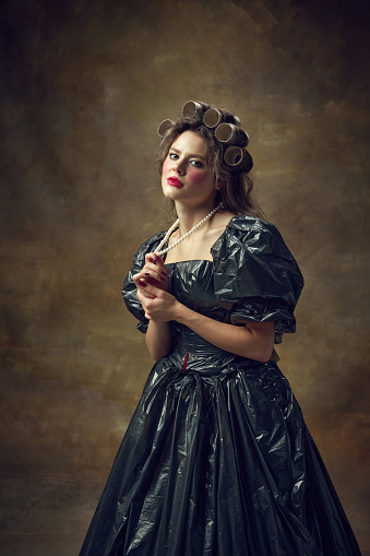 Princess. Portrait of woman dressed in paper tube wig and black dress made of garbage bags touching pearl necklace looking at camera. Concept of environmental issues, recycling, style, fashion.