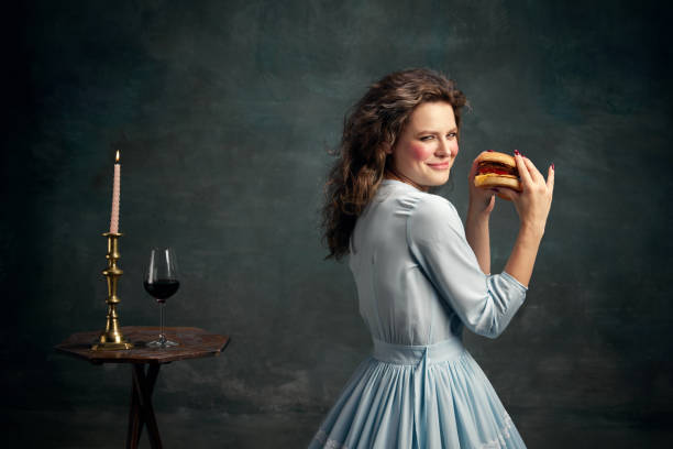 Princess. Portrait of aristocratic slyly looking woman wearing blue historical dress and holding hot big hamburger. Modern food art. Taste. Portrait of aristocratic slyly looking woman wearing blue historical dress and holding hot big hamburger. Modern food art. Concept of healthy lifestyle, diet, style, fashion. mirror women baroque style fashion stock pictures, royalty-free photos & images