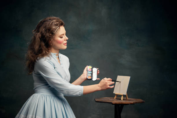 Artist. Portrait of charming aristocratic woman wearing blue historical dress holding eye shadow palette in hand and draws it on small canva. Artist. Portrait of charming aristocratic woman wearing blue historical dress holding eye shadow palette in hand and draws it on small canva. Concept of modern art, painting, style, fashion. mirror women baroque style fashion stock pictures, royalty-free photos & images