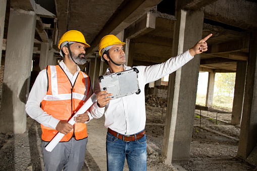 Two Indian male civil engineers or architect wearing helmet and vest holding paperwork blueprint at construction site discussing real estate project