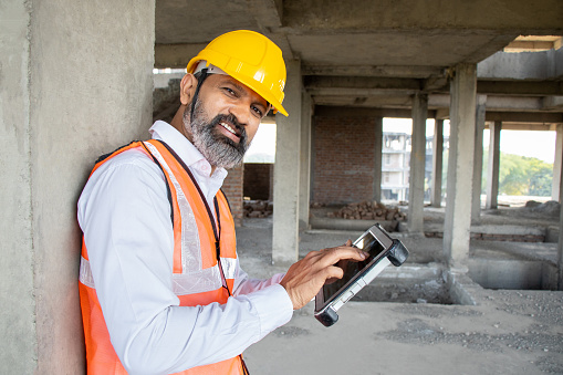 Mature Indian male civil engineer or architect wearing helmet and vest holding digital tablet blueprint at construction site.