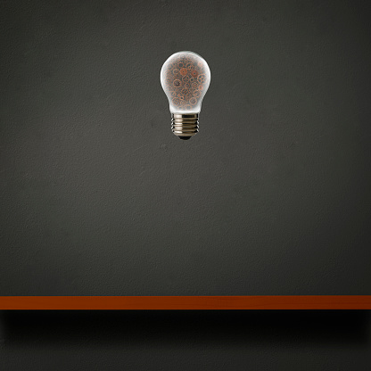 Floating a light bulb with metal gears in mid-air over the wooden shelf against dark concrete wall with copy space.