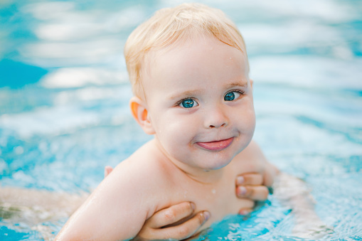 A baby swimming instructor holding and swimming with a baby during a class in Boldon, North East England. The baby is looking at the camera while the instructor looks at her and the baby's father watches and claps them in the background.