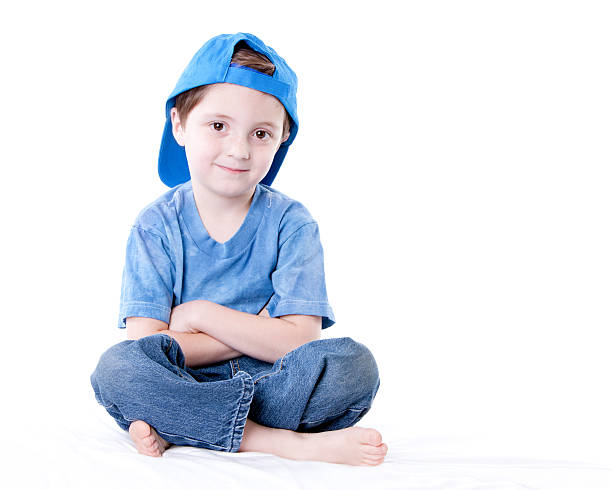 A full length image of a smiling caucasian little real boy sitting cross legged with his arms crossed, wearing a blue shirt and baseball cap.