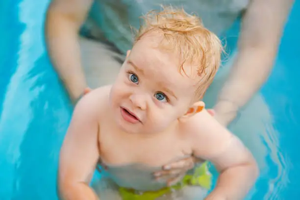 Photo of Portrait of small red-haired boy bathes in pool with hand support, baby swimming in water, summer leisure