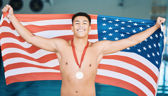 A group of four multi-ethnic children standing at the side of a swimming pool, holding American flags, smiling and looking at the camera.