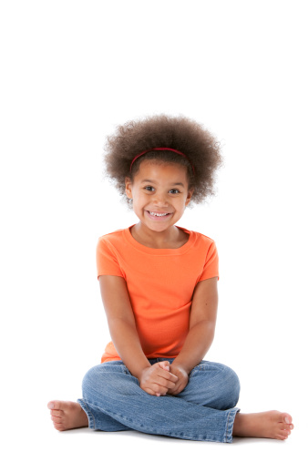 A happy, smiling ethnic (African American) little girl sits cross-legged on the floor with her hands in her lap as if in anticipation of something.
