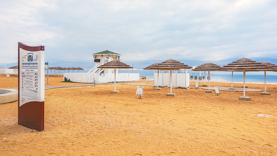 Ein Bokek, Israel - January 22, 2020: Empty Dead Sea sandy beach with sun umbrellas and info board, lifeguard hut near, calm water and overcast sky in distance
