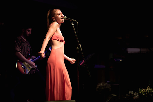 A striking on-stage portrait of a young woman in her early twenties with blonde hair and fair skin, emotionally singing into a microphone, set against a dark background