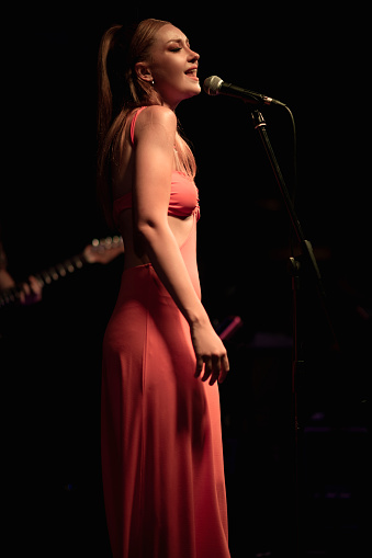 A striking on-stage portrait of a young woman in her early twenties with blonde hair and fair skin, emotionally singing into a microphone, set against a dark background