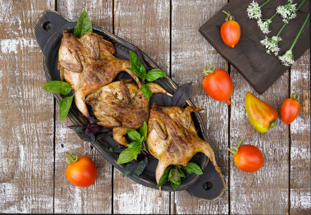 A wooden dish with grilled quails. Tomatoes and bell peppers are lying on a wooden table.