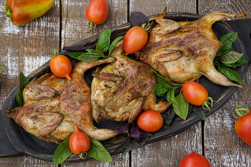 A wooden dish with grilled quails. Tomatoes and bell peppers are lying on a wooden table.