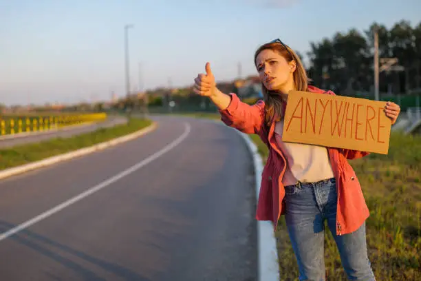 Woman is hitchhiking on roadside trying to stop car. She is holding cardboard with inscription.