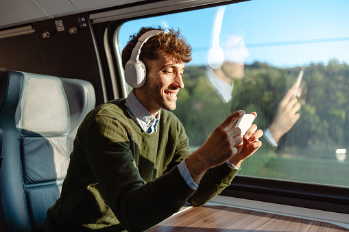 Portrait of a cheerful man in smart casual clothes watching something on smart phone while traveling by train. He is wearing wireless headphones connected to his smart phone.